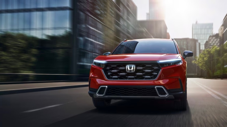 Honda CR-V 2025 rouge en vue avant roulant dans une rue urbaine bordée de bâtiments modernes.