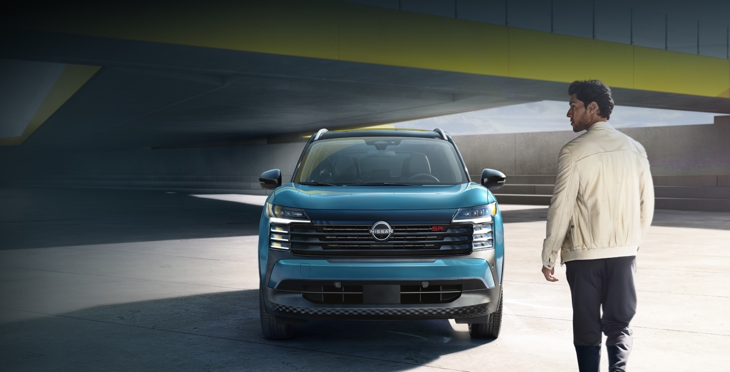 Vue avant complète du Nissan Kicks 2025 bleu sous un pont moderne, avec un homme marchant à proximité dans un décor urbain clair.